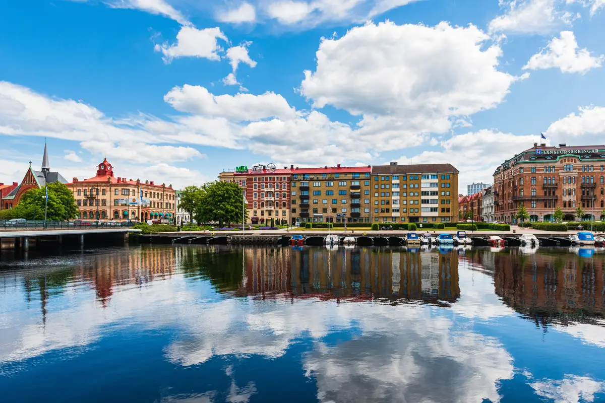Colorful buildings along the river in Halmstad, Sweden, reflecting on the calm water with a clear sky and boats docked at the riverside.