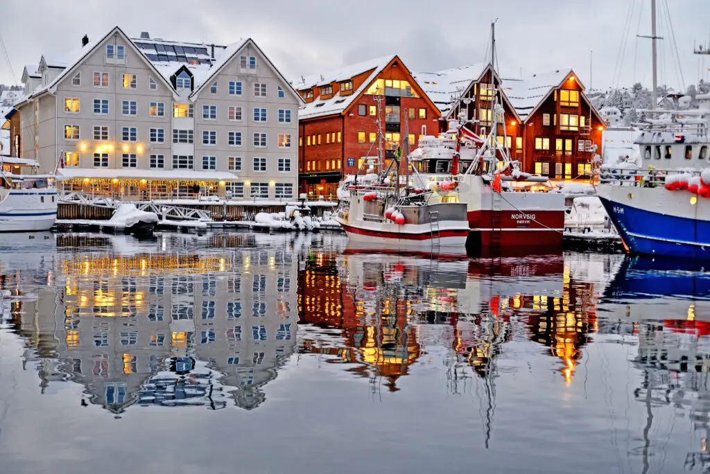 Winter evening view of Tromsø port in Norway, featuring colorful waterfront buildings and boats reflected in the water.