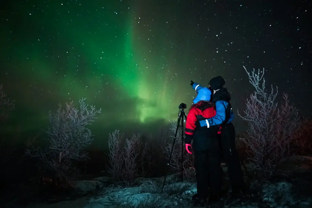 Two people observing the Aurora Borealis in Lapland, Finland, capturing the vibrant northern lights above a snowy landscape.