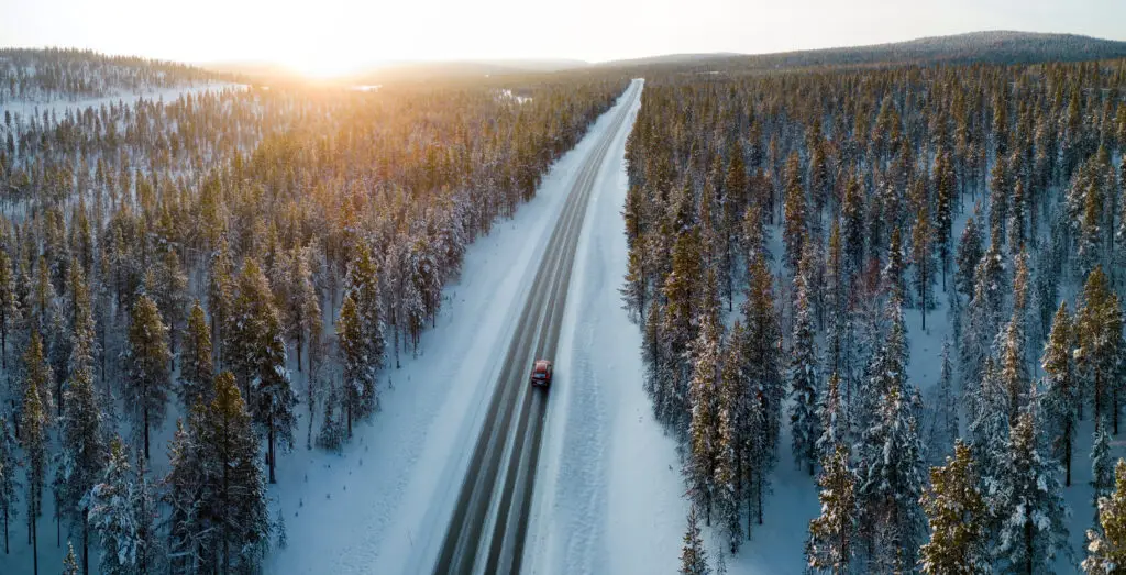Aerial view of a snow-covered road winding through a dense, frosty forest in Finnish Lapland during winter, showcasing the serene and scenic winter driving route.