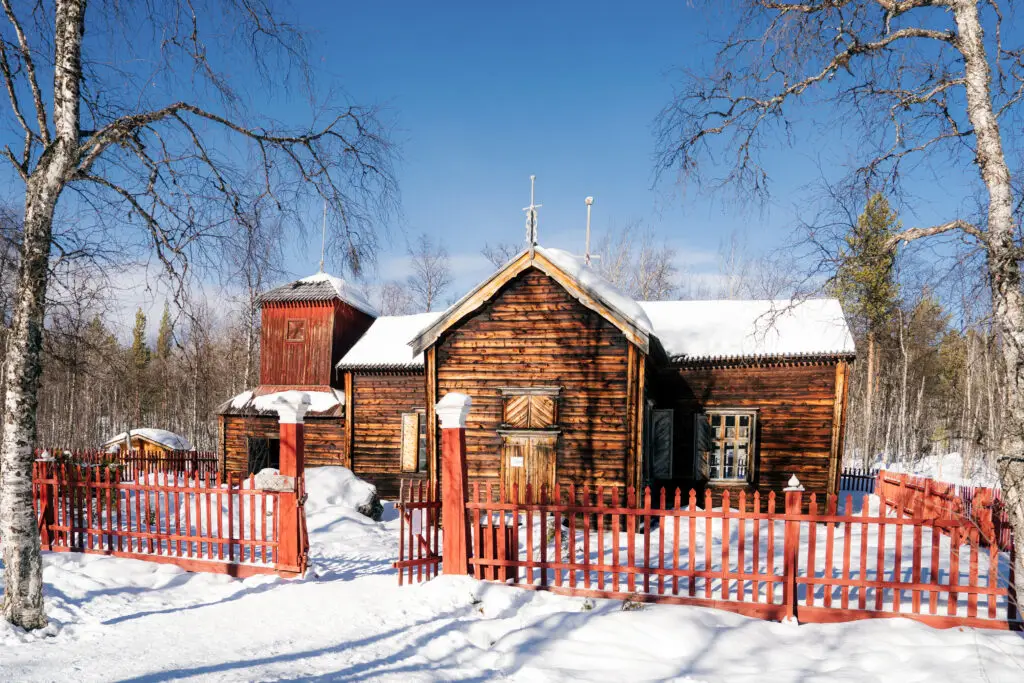 Traditional wooden Sámi church surrounded by tall birch trees and a red picket fence in Inari, Finnish Lapland, showcasing Finnish cultural heritage in a snowy landscape.