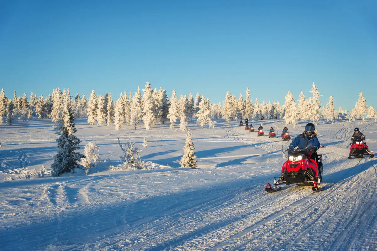 Tourists riding snowmobiles through the snowy landscape of Lapland near Saariselka, Finland, under a clear blue sky.