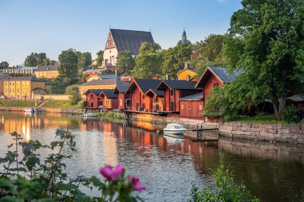 Summer evening view of Porvoo, Finland featuring the idyllic riverfront with traditional red wooden warehouses and a historic church in the background.