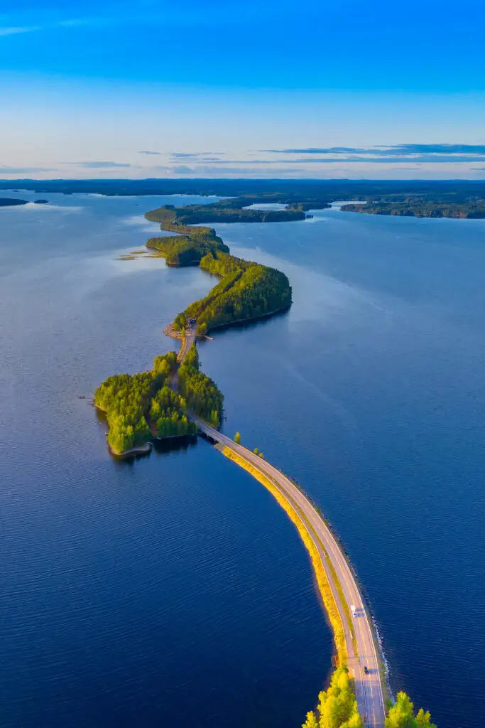 Aerial view of Pulkkilanharju Ridge in Paijanne National Park, Finland, showcasing a narrow road winding through vibrant green forests and deep blue lakes on a sunny summer day.
