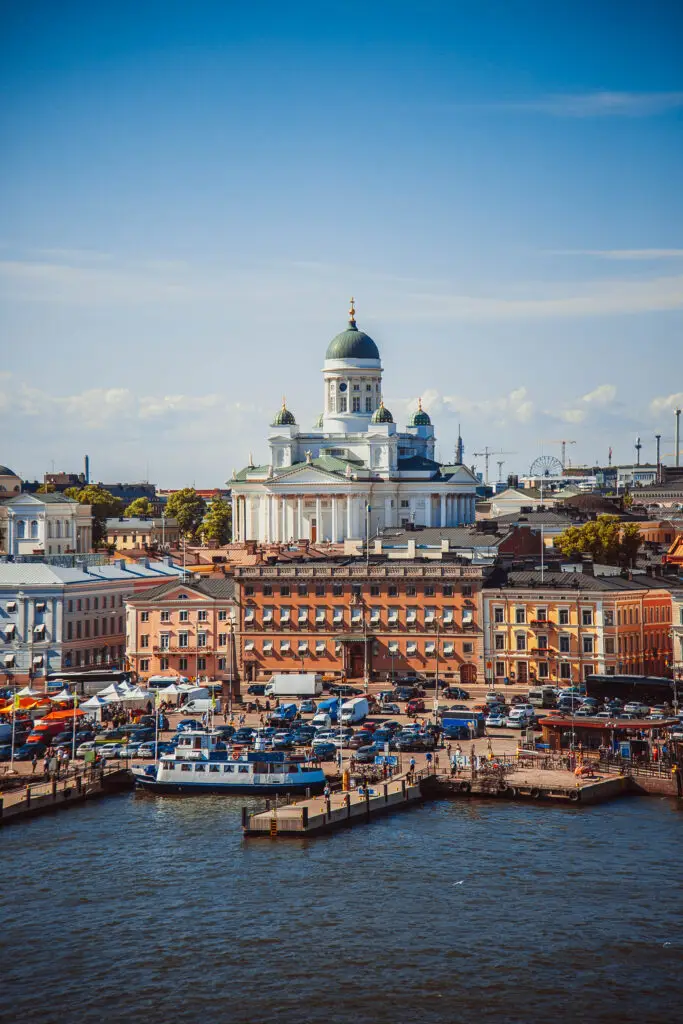 Summer panorama of Helsinki showcasing the Helsinki Cathedral and bustling market square filled with tourists and boats docked at the harbor.