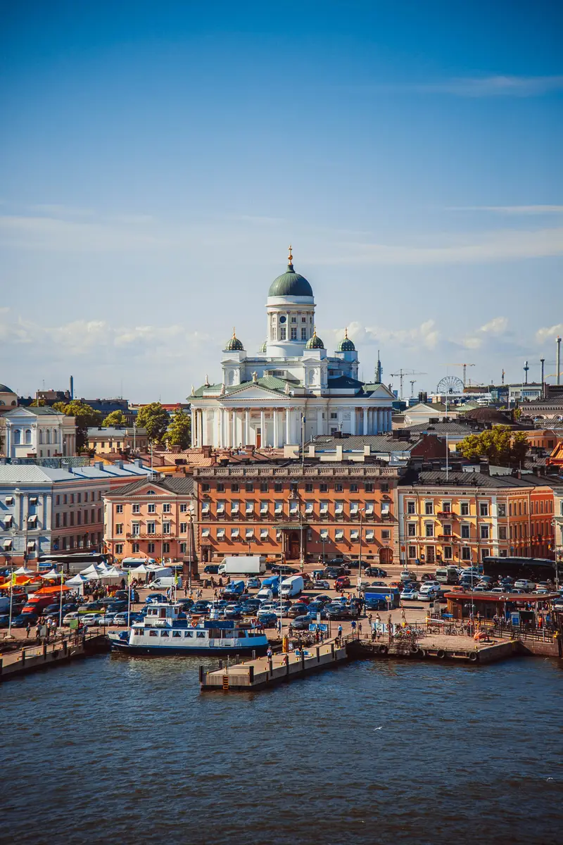 Summer panorama of Helsinki showcasing the Helsinki Cathedral and bustling market square filled with tourists and boats docked at the harbor.