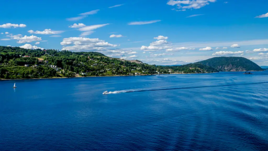 Panoramic view of Oslofjord featuring a speedboat and a sailboat, with rolling green hills and residential areas near Sætre, Norway.