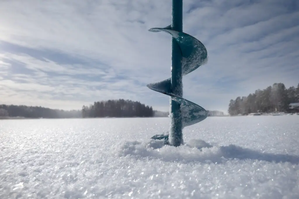 Ice auger drilling into a frozen lake with snowy landscape under a clear blue sky in Finland, depicting winter fishing activities.