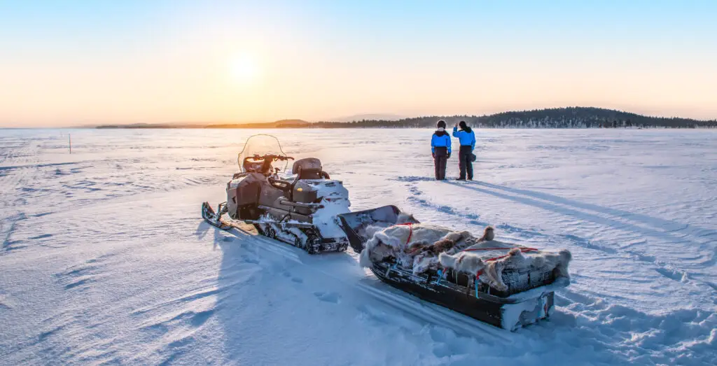 Young couple admiring the sunrise near a snowmobile on the frozen Lake Inari, Lapland, Finland, showcasing winter tourism and adventure in the Arctic.