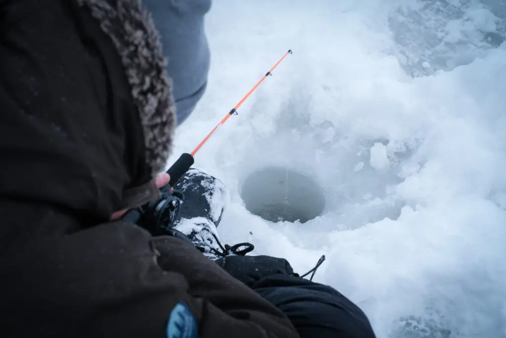 Person ice fishing in Finland, sitting by a drilled hole in the frozen lake with a fishing rod, showcasing a popular Finnish winter activity
