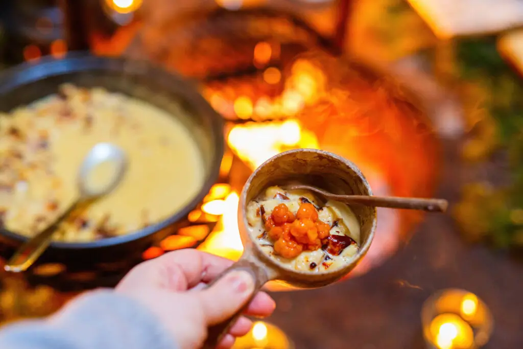 Hand holding a traditional Lappish baked cheese topped with cloudberry jam in a wooden kuksa cup, with a cozy, fire-lit setting in the background, showcasing Scandinavian culinary culture.
