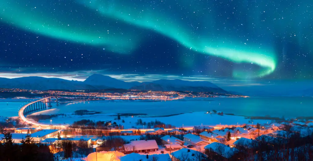 Stunning view of the Northern Lights over Tromso with Sandnessundet Bridge illuminated at night, showcasing a snowy landscape and vibrant city lights in Norway.